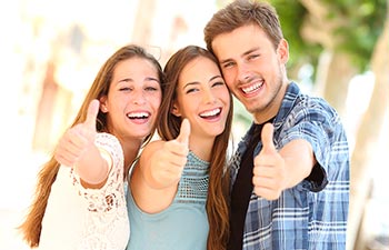 Two young women and one man with perfect smiles showing their thumbs up.