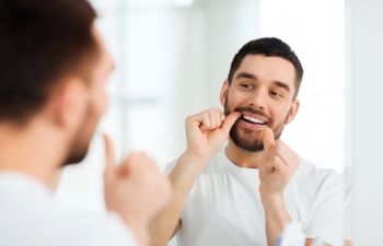 man with dental floss cleaning teeth in front of the bathroom mirror