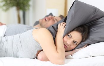 woman having trouble sleeping and covering her ears with a pillow due to her husband's loud snoring